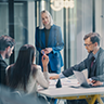Four people are in a modern office meeting. Three are seated at a table with laptops and papers, while one person stands, speaking to the group. The atmosphere appears professional and collaborative. EFESO Management Consultants